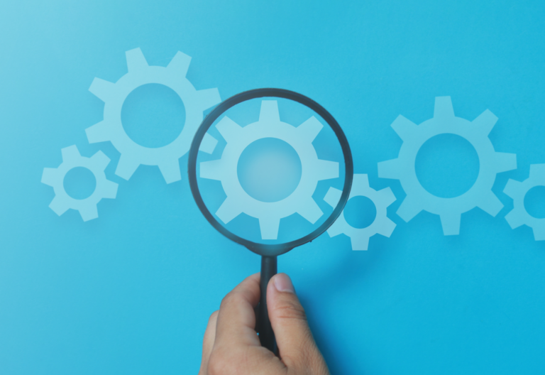 Eine Hand hält eine Lupe über Zahnräder auf blauem Hintergrund.